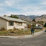 Two people walk along a sidewalk in front of a beige ranch-style house with a 'For Sale' sign; a quiet street with parked cars and distant mountains.
